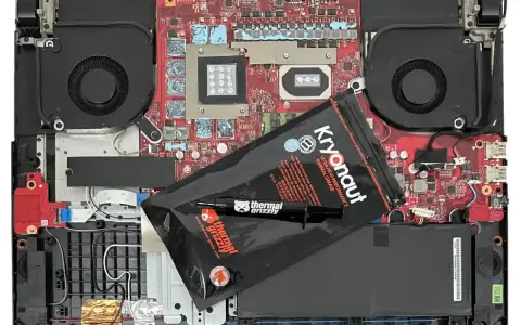 Wnętrze laptopa z wentylatorami i płytą główną, gotowe do wymiany pasty termoprzewodzącej. Opakowanie Kryonaut Thermal Grizzly leży na komponentach.