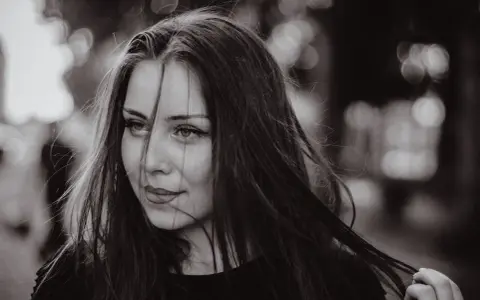 Portret młodej kobiety z burzą ciemnych włosów. Jej spojrzenie jest lekko zadumane, a usta subtelnie uśmiechnięte. Grzywka dodaje jej tajemniczości.