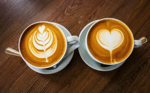 Zwei Cappuccinos mit Latte Art auf einem Holztisch. Sie bestehen aus Espresso, heißer Milch und Milchschaum.