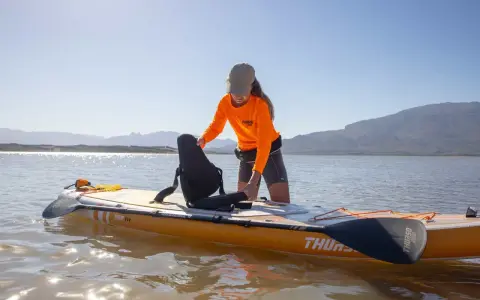Frau richtet Sitz auf SUP-Board ein, das wie ein Kajak genutzt werden kann. Sonnenschein, Berge und See im Hintergrund.