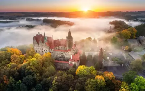 Zamek Książ otoczony jesiennymi drzewami, wyłaniający się z porannej mgły. Słońce wschodzi nad wzgórzami, malując niebo na pomarańczowo.