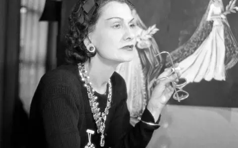 Coco Chanel, icona di stile, con occhiali in mano, sullo sfondo schizzi di abiti da sposa. Un'immagine che racconta la sua storia.