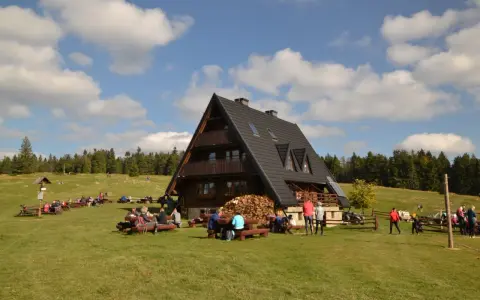 Drewniana bacówka z ludźmi siedzącymi przy stołach na trawie, w tle las i niebo z chmurami.
