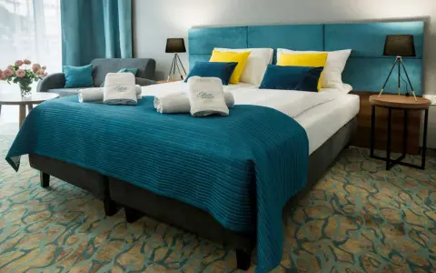 Przytulny pokój hotelowy z dużym łóżkiem, ozdobionym turkusową narzutą i żółto-niebieskimi poduszkami. Ręczniki z logo Stella Resort & Spa zachęcają do relaksu.