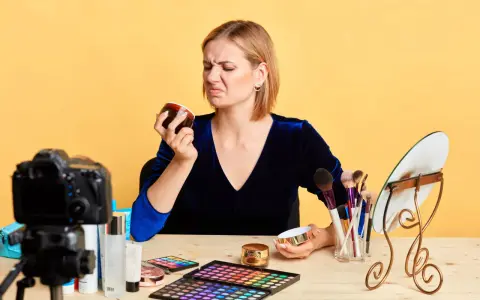 Donna con espressione disgustata annusa una crema viso chiusa, forse per verificarne la scadenza. Sul tavolo, trucchi e una telecamera.