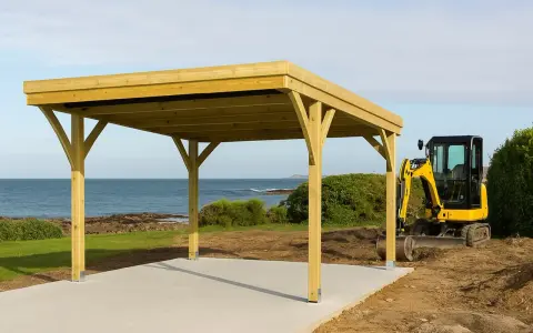 Un carport en bois est en cours de construction près de la mer. Une mini-pelle jaune est garée à côté, prête pour la suite des travaux. Une déclaration de carport est nécessaire pour ce projet.