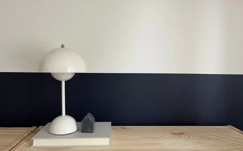 Lampe blanche sur un livre, devant un mur deux couleurs différentes, blanc et bleu nuit. Un petit objet gris en forme de maison est posé sur le livre.