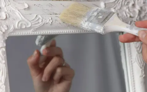 Pintura a la tiza blanco antiguo en un marco de espejo ornamentado. Una mano aplica la pintura con una brocha, creando un acabado desgastado.