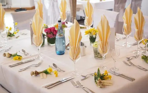 Elegancko nakryty stół weselny z żółtymi serwetkami, kwiatami i butelką wody. Każde miejsce ma imienną wizytówkę, jak dla drużby.