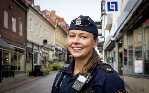 En ung poliskvinna ler mot kameran på en gata i Sverige. Hon bär uniform och utrustning, redo att rycka ut om larmet går.