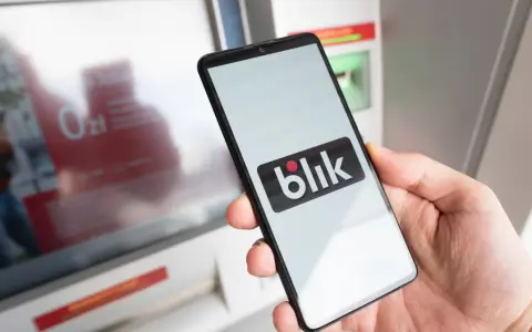 Dłoń trzyma smartfon z logo BLIK przed bankomatem.