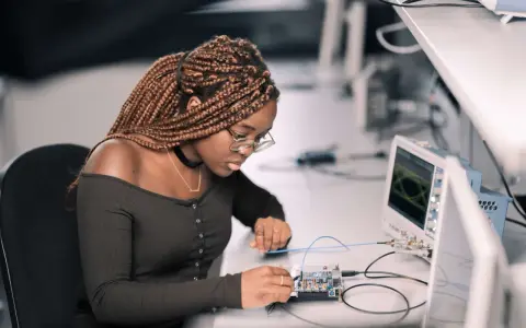 Junge Frau mit geflochtenen Haaren arbeitet konzentriert an einer Platine, während ein Oszilloskop im Hintergrund läuft. Ein spannendes elektrotechnik studium.