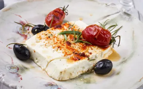 Feta al forno con pomodorini arrostiti, olive nere e rosmarino su un piatto rustico.