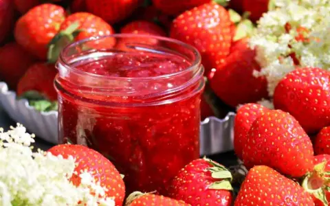 En burk med hemgjord jordgubbssylt omgiven av färska jordgubbar och blommor. Perfekt för ett enkelt jordgubbssylt recept.