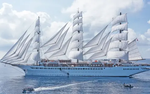 Ein majestätisches Segelschiff mit vollen weißen Segeln, das wie ein **sea cloud spirit** über das blaue Meer gleitet. Kleine Beiboote sind in der Nähe.