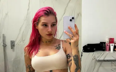 Młoda Leosia z kolorowymi włosami i licznymi tatuażami na ciele robi sobie selfie w łazience.