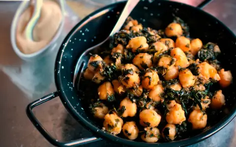 Un plato de garbanzos con espinacas, servido en una sartén negra con un tenedor. Al fondo, una pequeña taza con una salsa cremosa.