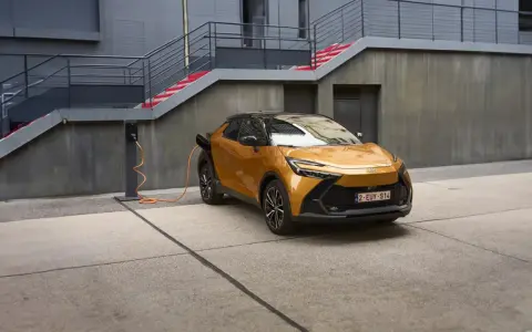 Złoty Toyota plug-in z pomarańczowym kablem ładowania podłączonym do stacji. Nowoczesna architektura w tle.