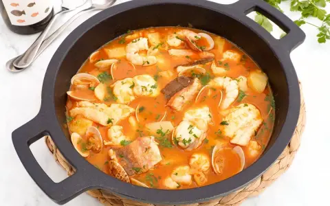 Una deliciosa cazuela de pescado con gambas, almejas y trozos de pescado blanco en una rica salsa roja, adornada con perejil fresco.