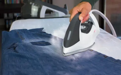 Hand using a steam iron on a denim shirt on an ironing board.