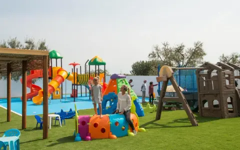 Un parco giochi colorato in una masseria per bambini in Puglia, con scivoli, tunnel e altalene. Famiglie si divertono sotto il sole.