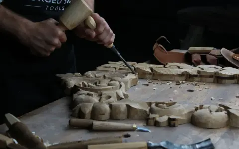 Händer formar träsnideri med mejsel och klubba. Det ser ut som att träet är mjukt som lera, perfekt för detaljerat arbete.