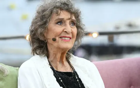 Siv Malmkvist, med sitt karaktäristiska lockiga gråa hår och ett varmt leende, sitter och tittar åt sidan. Hon bär en vit kofta över en svart klänning och smycken.