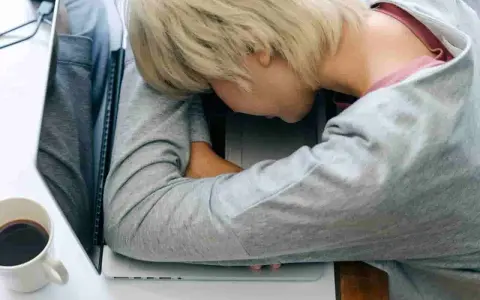 Zmęczona osoba śpi na laptopie. Czy leczenie odwykowe a zwolnienie z pracy to trudny wybór? Kawa obok.