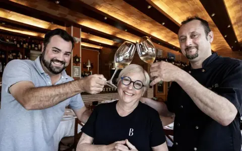 Dos hombres y una mujer brindan con copas de vino en Ca la Pepa, un restaurante acogedor con estanterías de vino.