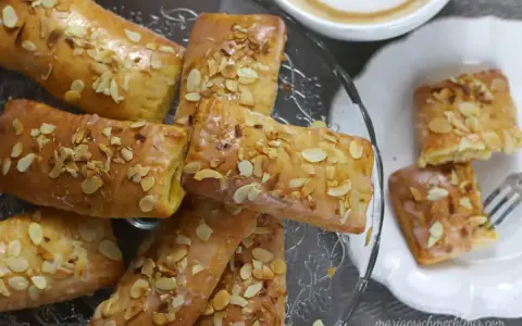 Omas Apfeltaschen Rezept: goldbraune, glasierte Gebäckstücke mit Mandelblättchen, serviert auf einem Glasständer und einem Teller, daneben eine Tasse Kaffee.