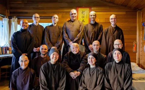 A group of monks and nuns, including an elder monk person in the center, pose for a photo in a room with wooden walls.