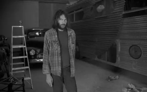 Neil Young, jung und nachdenklich, steht vor einem umgebauten Bus. Eine Leiter und ein Oldtimer im Hintergrund.