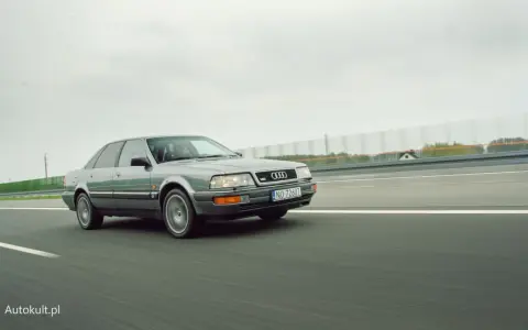Szary Audi V8 pędzi po autostradzie, chmury na niebie i zielone drzewa w tle.