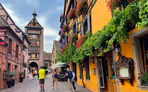 Cobblestone street in a picturesque Alsace town, with colorful half-timbered buildings adorned with flowers. A clock tower stands tall in the background.