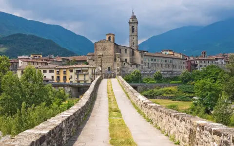 Ponte medievale in pietra che conduce a un borgo con chiesa e campanile, incastonato tra le verdi montagne degli Appennini Emilia Romagna.