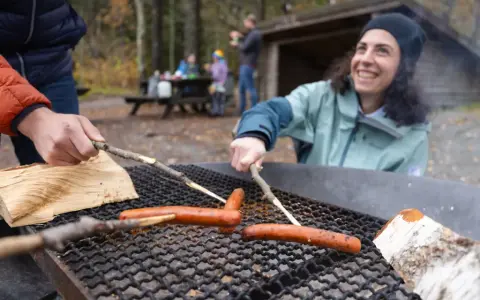 Vänner grillar korv vid en eldstad på Otto Pers gård. En kvinna ler glatt medan hon grillar.