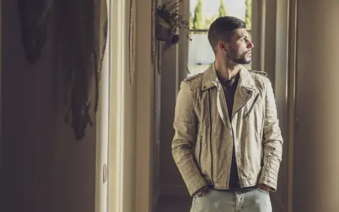 Uomo con un outfit giacca pelle uomo color crema, jeans chiari, in un corridoio luminoso.