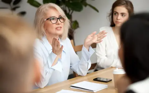 Kobieta w okularach, omawiająca ocenę pracy nauczyciela, gestykuluje do grupy.