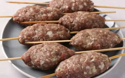 Siedem surowych kofta kebab na patyczkach, ułożonych na szarym talerzu, gotowych do grillowania.