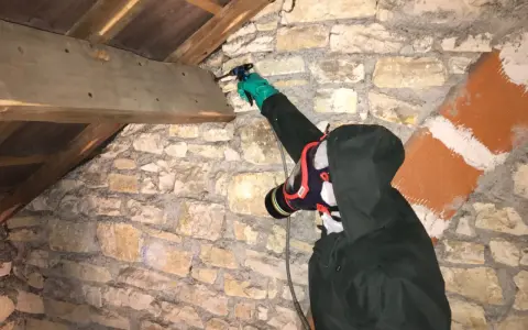 Personne en combinaison et masque effectuant un traitement de charpente par pulvérisation dans un grenier en pierre.