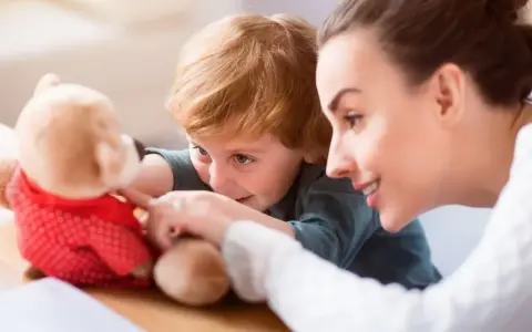 Uśmiechnięta opiekunka do dziecka bawi się z chłopcem pluszowym misiem.
