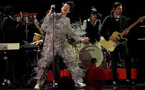 Harry Styles i glittrig outfit på en magisk harry styles konsert. Bandet spelar bakom honom, en gitarrist i kostym och en trummis.