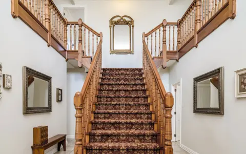 Elegante escalera antigua con alfombra de estilo persa y barandillas de madera. Un espejo dorado decora la pared central, complementando el revestir escalera antigua.