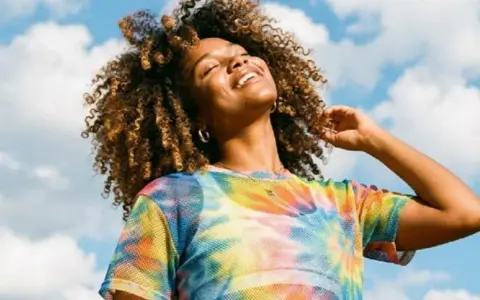 Une femme souriante aux cheveux bouclés, sous un ciel bleu nuageux. Elle porte un t-shirt tie-dye coloré. Une belle image pour apprendre comment rendre une photo flou.