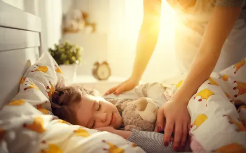 Mama pomaga dziecku zasnąć, kładąc rękę na jego ramieniu. Słodki sen malucha to pierwszy krok, jak nauczyć dziecko zasypiać samo.