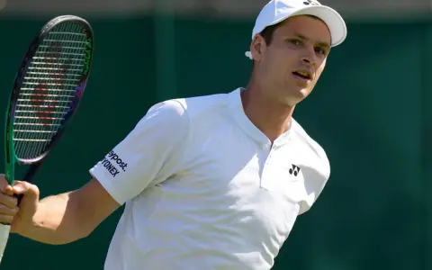 Hubert Hurkacz w białej koszulce i czapce, gotowy do uderzenia rakietą na Wimbledonie.
