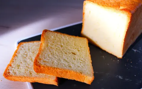 Pain de mie moelleux, parfait pour une recette pain de mie boulangerie maison. Deux tranches dorées et un pain entier sur un plateau sombre.