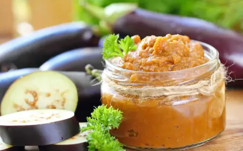 Un bocal de le vrai caviar d'aubergine, garni de persil frais, avec des aubergines entières et tranchées en arrière-plan.