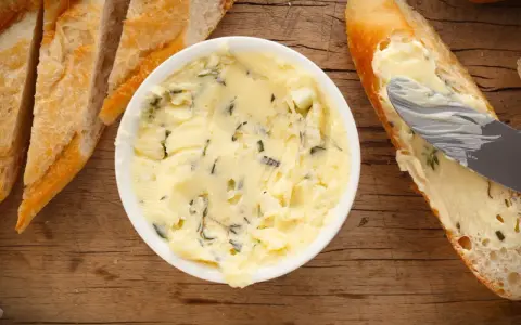 Knoblauchbutter selber machen ist einfach! Hier ist eine Schüssel mit selbstgemachter Knoblauchbutter, daneben Baguettescheiben und ein Messer, das Butter auf ein Brot streicht.