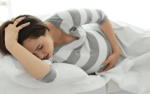 Schwangere Frau liegt auf dem Bett, hält sich den Kopf und fragt sich: "Vorwehen, wie lange noch bis zur Geburt?
